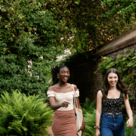 Two smiling students walk side by side in a leafy green garden along side a brick wall