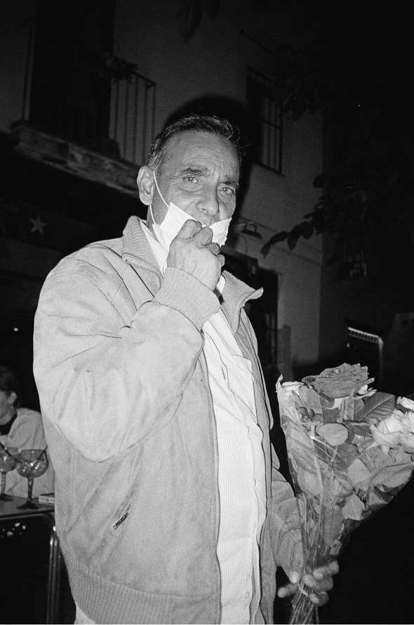 A black and white night-time photo of an elderly man holding a bunch of roses and wearing a face mask